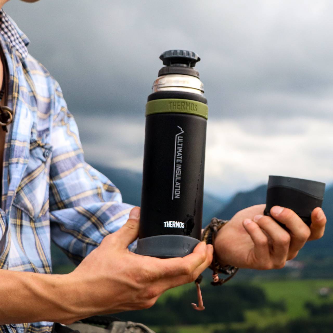 Eine Person hält eine schwarze Thermosflasche mit einer olivgrünen Kappe und den abgenommenen Deckel in den Händen. Im Hintergrund ist eine verschwommene Landschaft mit grünen Wiesen und Hügeln unter einem wolkigen Himmel.