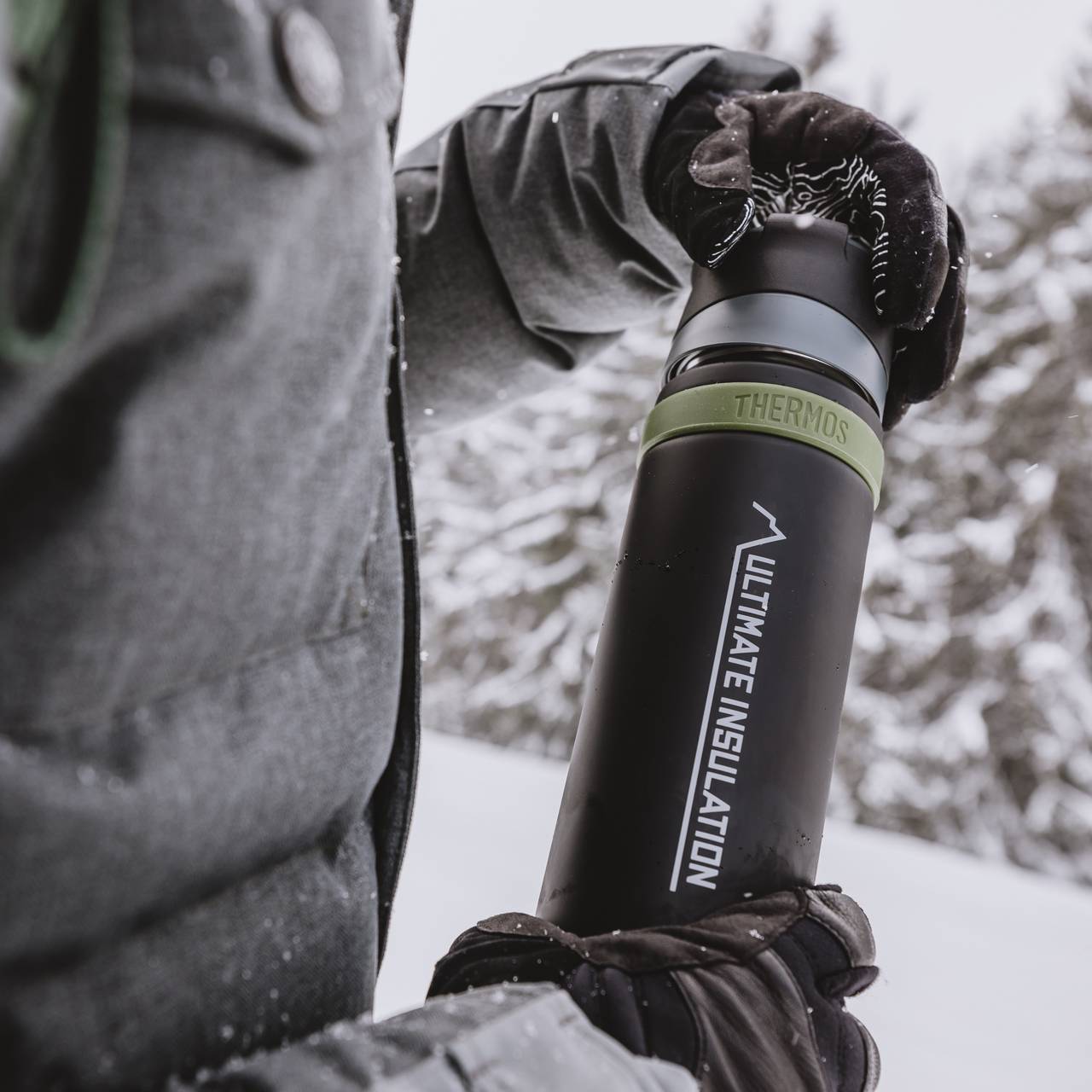 Eine Person in warmer Winterkleidung hält eine schwarze Thermoskanne mit grünem Deckel, auf der "Ultimate Insulation" steht. Im Hintergrund sind schneebedeckte Bäume zu sehen. Die Szene vermittelt eine kalte, winterliche Umgebung.