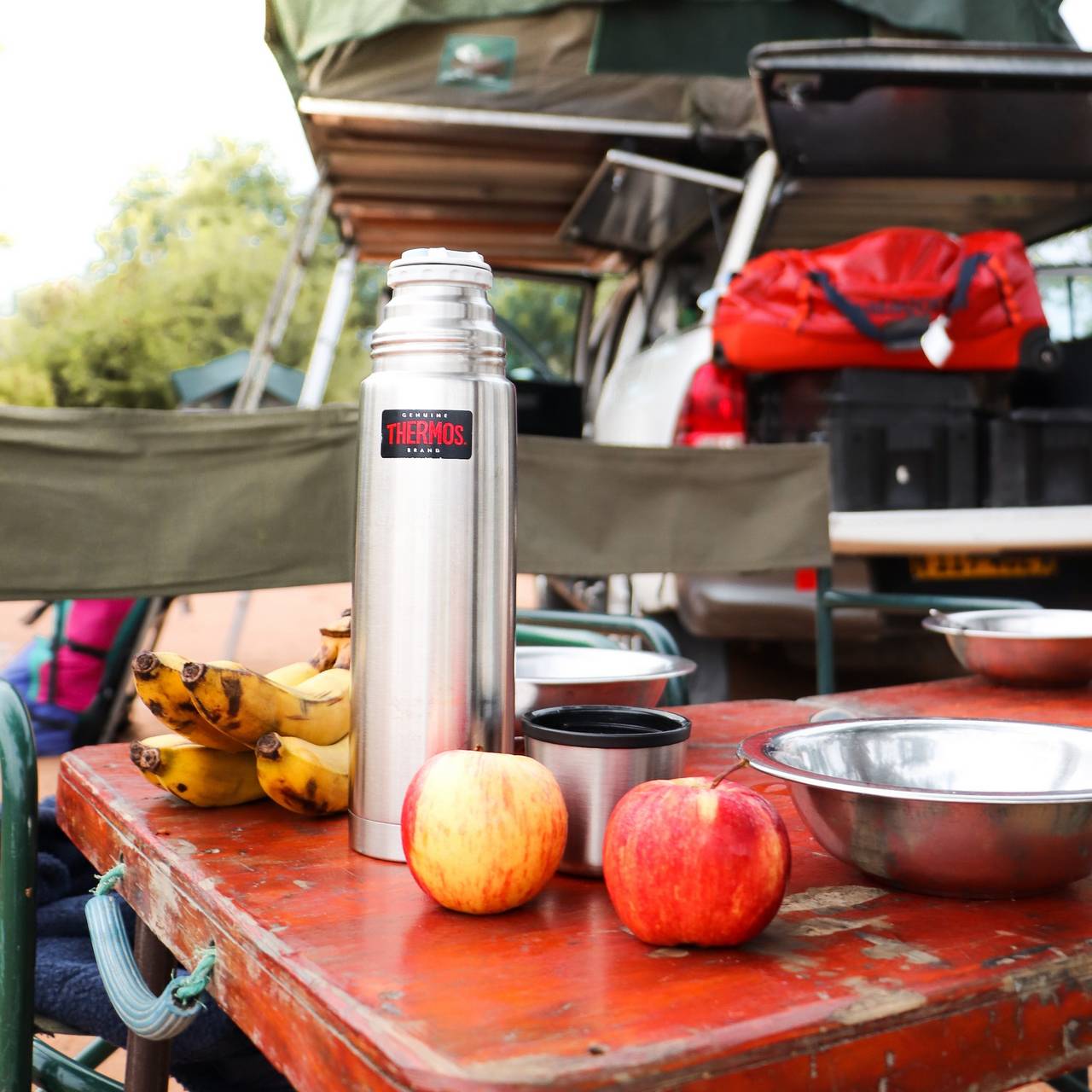 Auf einem Tisch steht eine silberne Thermoskanne neben zwei Äpfeln und Bananen. Im Hintergrund sind Campingstühle und ein Fahrzeug mit Taschen. Es vermittelt eine Outdoor-Atmosphäre.