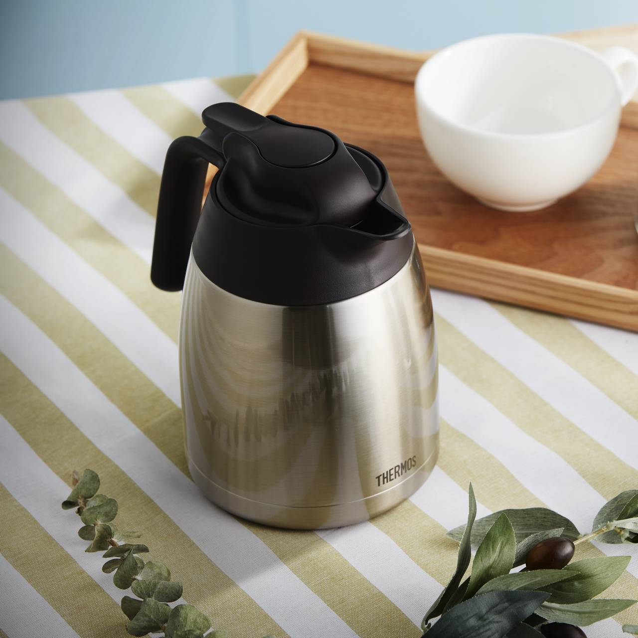 Eine silberne Thermoskanne mit schwarzem Deckel steht auf einer gestreiften Tischdecke. Im Hintergrund befindet sich ein Holztablett mit einer weißen Tasse darauf, sowie einige grüne Blätter als Dekoration.