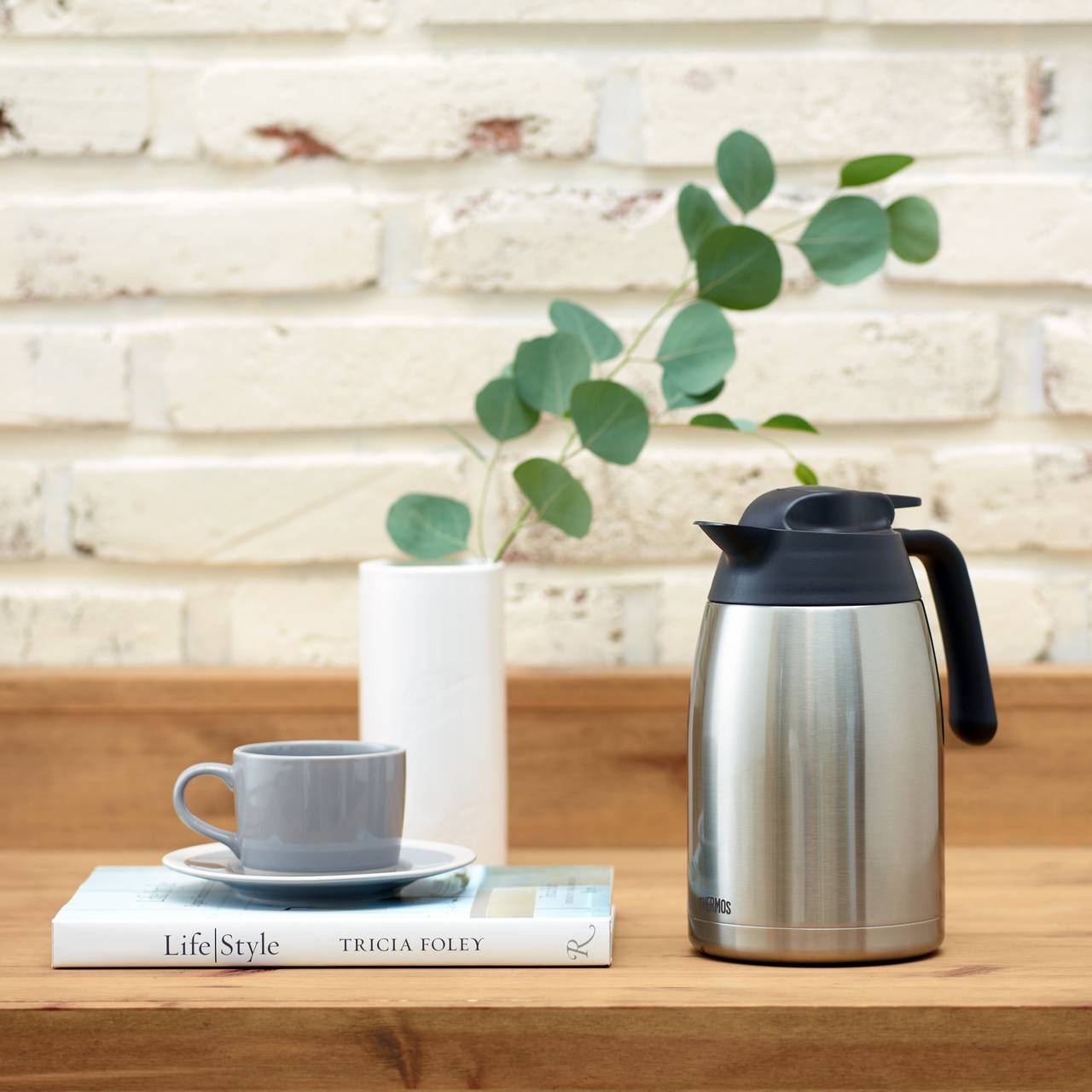 Ein Tisch mit einer silbernen Thermoskanne und einer grauen Tasse auf einem Buch. Daneben steht eine Vase mit Eukalyptusblättern. Der Hintergrund ist eine weiße Ziegelwand.