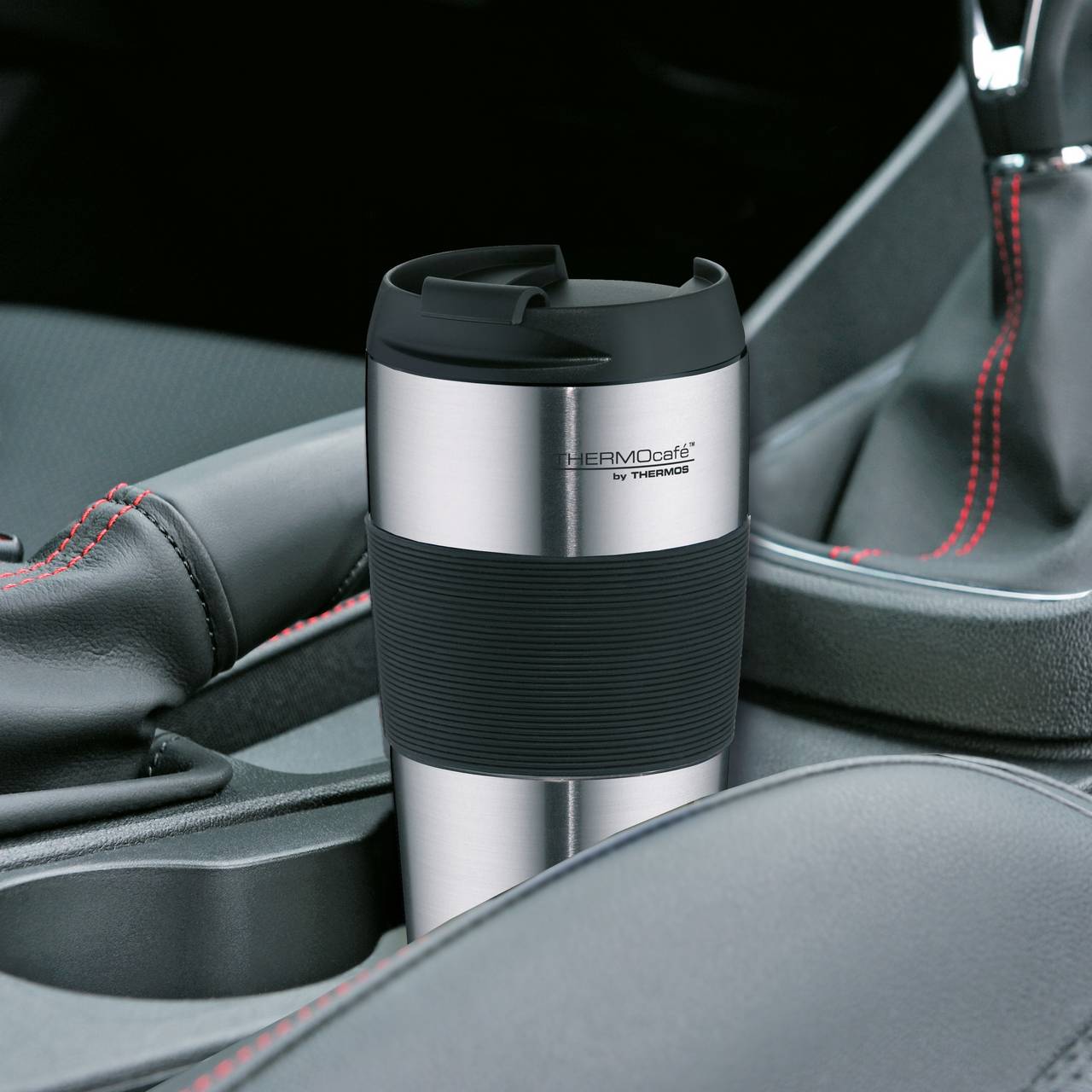 Ein silberner Thermobecher mit schwarzem Griff steht in einem Getränkehalter in einem Auto. Um den Getränkehalter herum sind schwarze Autositze und schwarze Nähte mit roten Akzenten zu sehen. Der Becher trägt die Aufschrift "THERMOcafé by THERMOS".