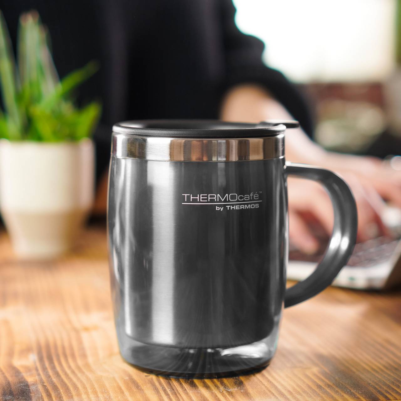Auf dem Bild ist ein metallener Thermosbecher mit Henkel auf einem Holztisch zu sehen, auf dem "THERMOcafé by THERMOS" steht. Im Hintergrund ist eine unscharf gehaltene Person, die an einem Laptop arbeitet, sowie eine Pflanze zu erkennen.