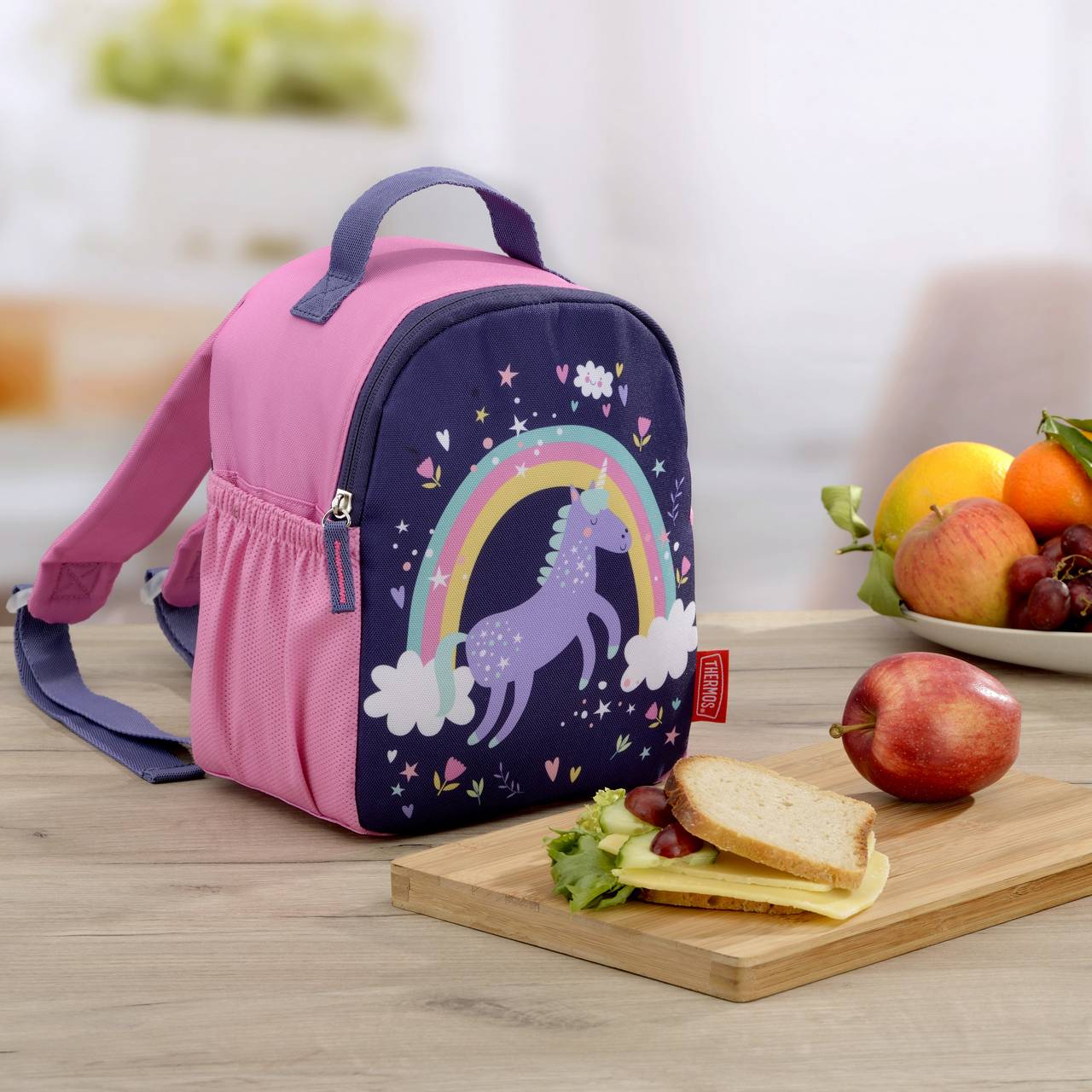 Auf einem Holztisch steht ein bunter Kinderrucksack mit Einhornmotiv und Regenbogen. Daneben liegt ein Brett mit einem belegten Sandwich und einem Apfel, im Hintergrund eine Schale mit Obst.