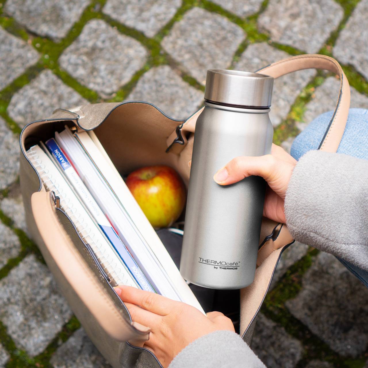 Eine Person hält eine silberne Thermosflasche und hat eine offene Handtasche auf Kopfsteinpflaster mit Moos. In der Tasche sind Notizbücher und ein Apfel.