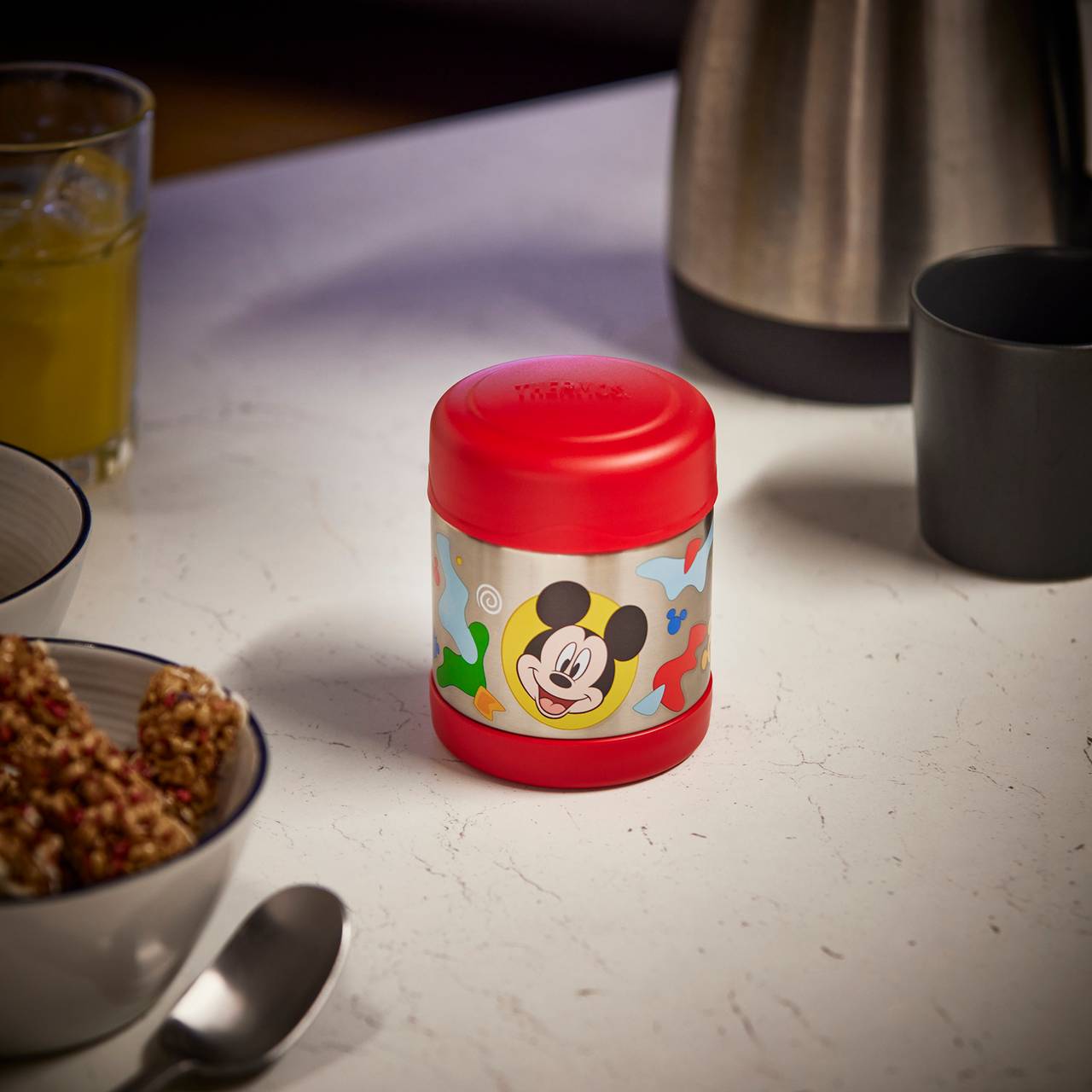 Ein roter Thermobehälter mit einem Cartoon-Gesicht steht auf einem Tisch. Daneben sind eine Tasse, eine Thermobehälter und eine Schale mit Getreidestücken. Im Hintergrund steht ein Glas mit gelbem Getränk.