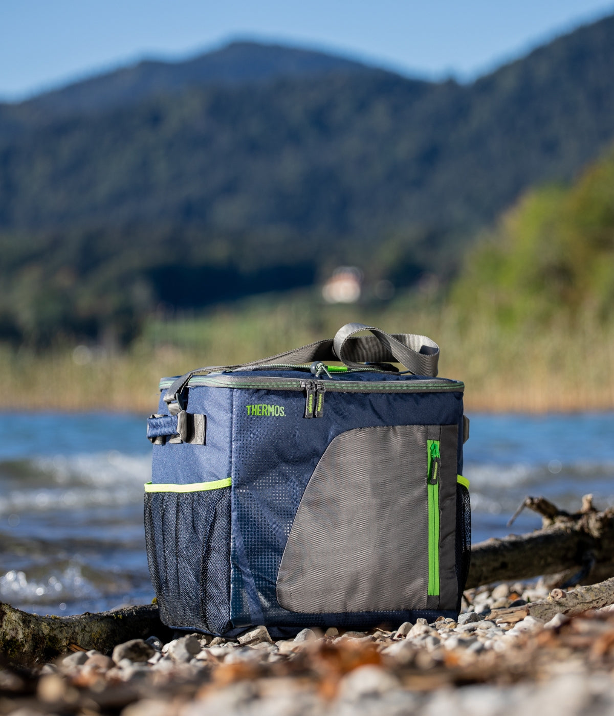 Eine blaue Radiance Kühltasche steht an einem steinigen Seeufer vor Bergen.