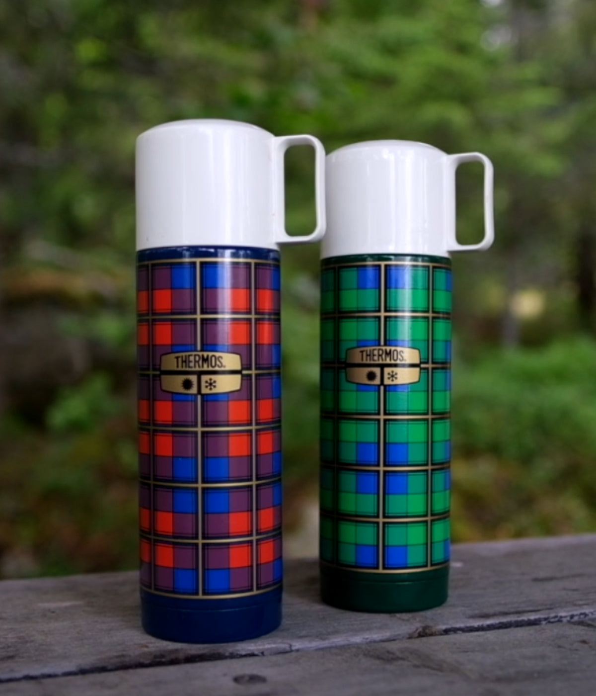 Zwei karierte Revival Thermosflaschen in Rot-Blau und Grün-Blau stehen auf einem Holztisch im Grünen.