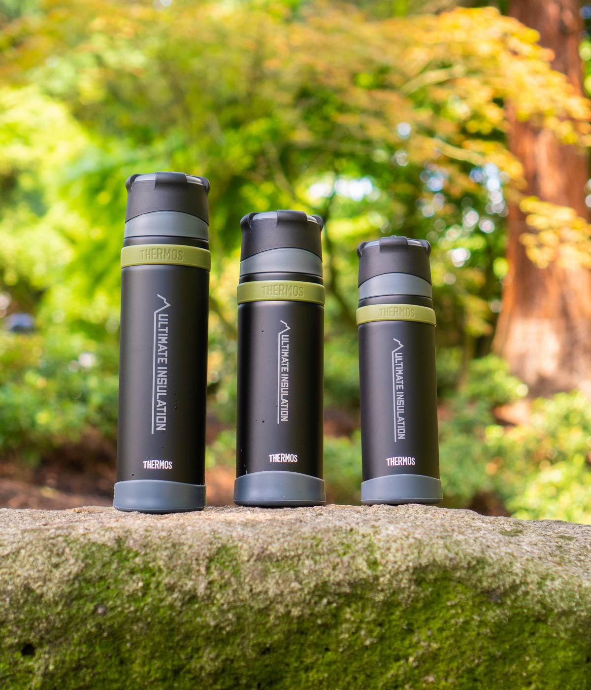 Drei dunkle THERMOS Mountain Flaschen stehen auf einem moosigen Felsen vor einem Waldhintergrund.