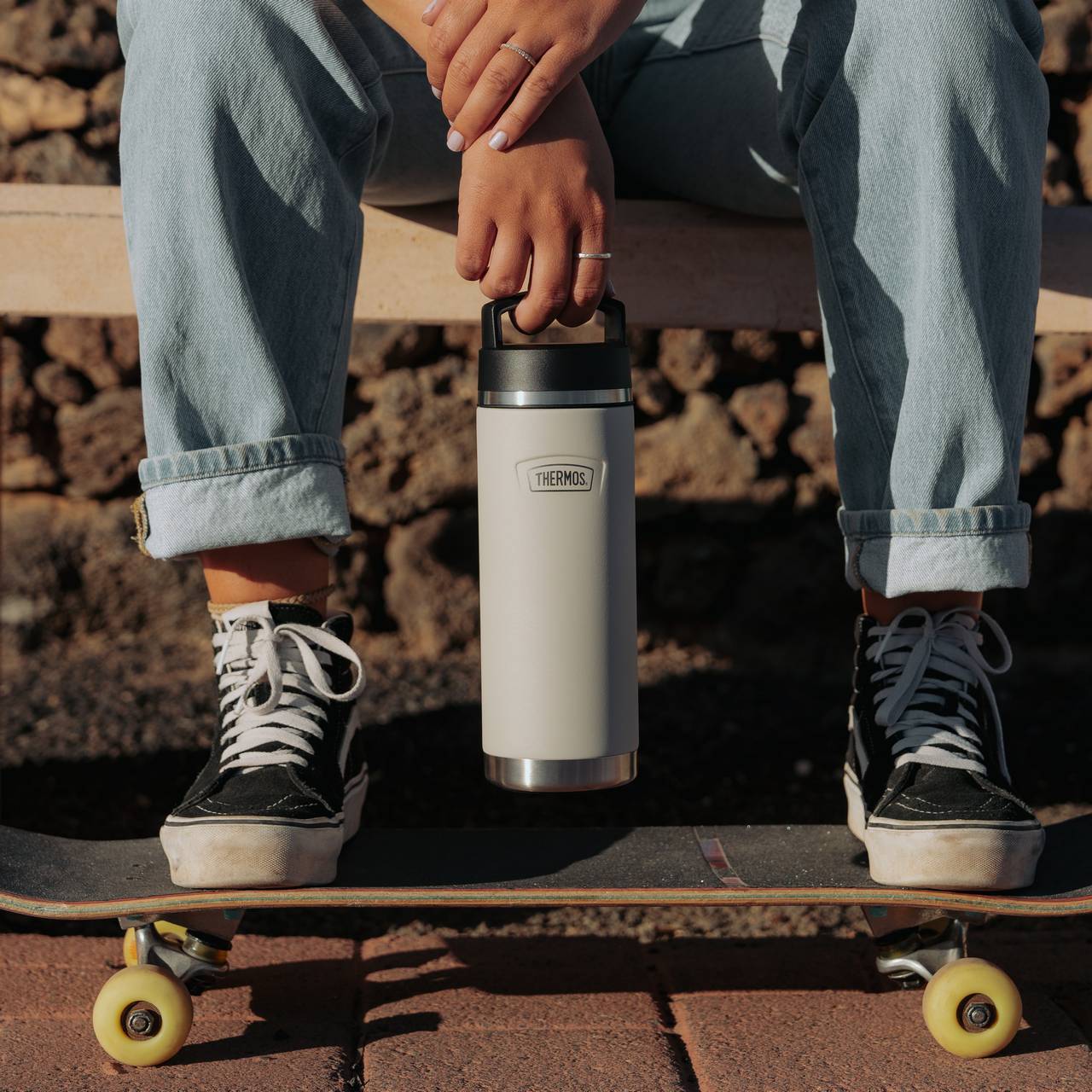 Eine Person sitzt auf einer Bank, hält eine Thermosflasche und hat ihre Füße auf ein Skateboard mit gelben Rädern gestellt. Sie trägt blau Jeans und schwarze Sneakers. Der Hintergrund zeigt eine Steinmauer und das Bild ist bei Tageslicht aufgenommen.