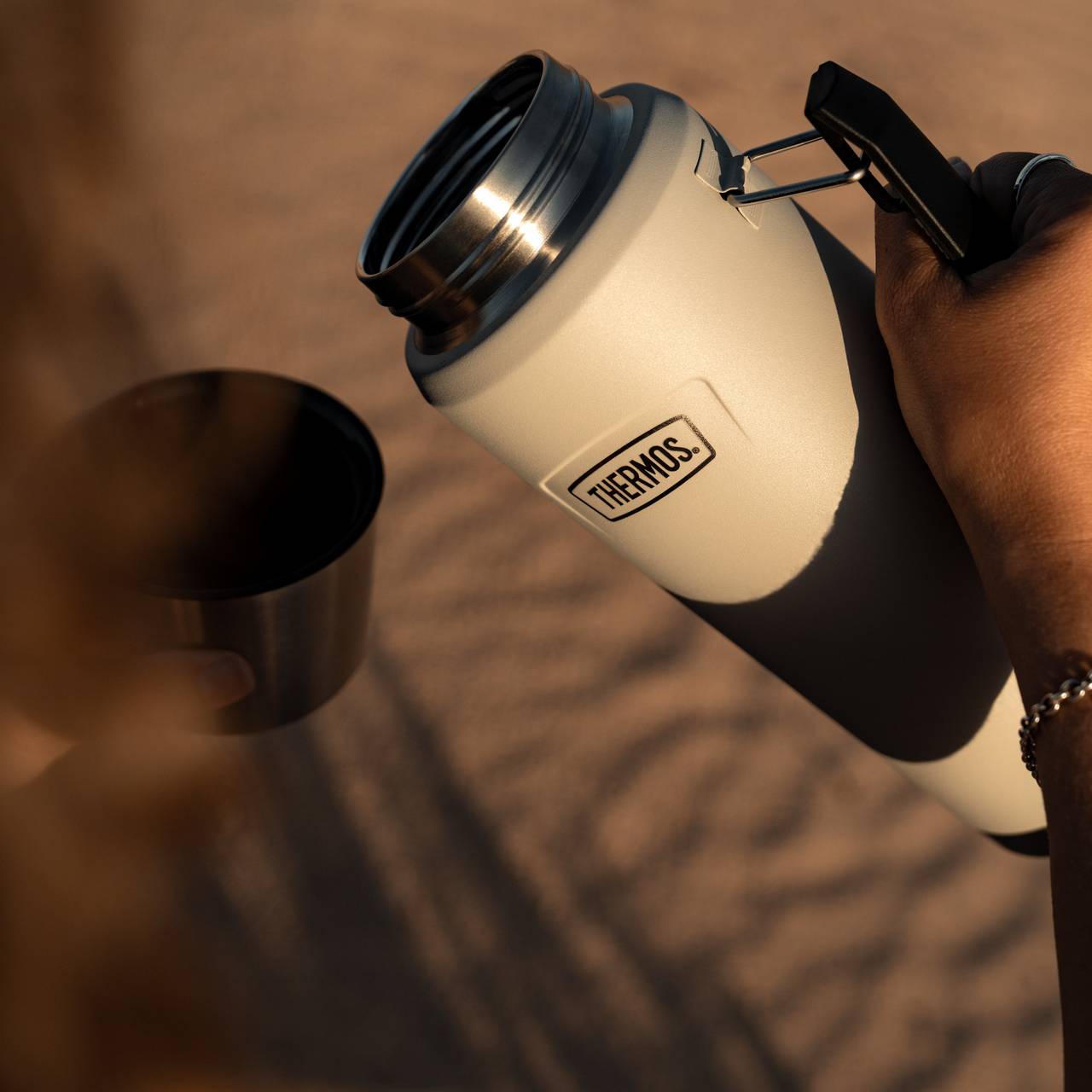 Eine Person hält eine beige Thermoskanne mit geöffnetem Deckel über einem kleinen, dunkel gefärbten Becher. Im Hintergrund ist sandiger Boden leicht verschwommen sichtbar, was auf eine sonnige Outdoor-Umgebung hindeutet.